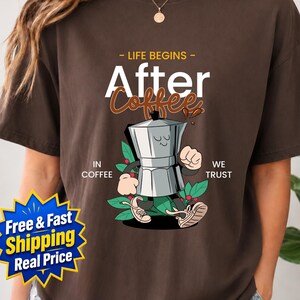 May include: A dark brown short-sleeve t-shirt featuring a graphic of a cartoon moka pot with arms and legs. The text reads "LIFE BEGINS After Coffee" and "IN COFFEE WE TRUST" in white and brown lettering, surrounded by green coffee leaves.