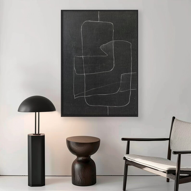 May include: A black and white abstract art print with a simple line design. The print is framed in a black frame and is hanging on a white wall. A black lamp with a round shade and a dark wood side table are also in the image.