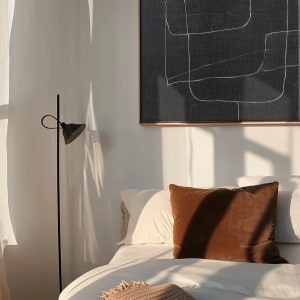 May include: A white bed with a brown throw pillow and a black floor lamp with a white shade. The bed is against a white wall with a black and white abstract painting hanging above it.