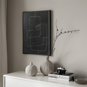 May include: A black and white abstract art print with a geometric design hangs on a light gray wall above a white table. Two ceramic vases, one gray and one white, are on the table with a vintage camera and a stack of books.