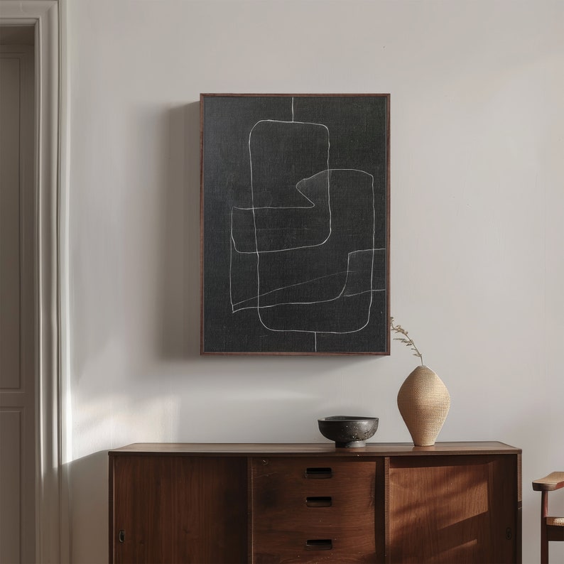 May include: A black and white abstract art print with a wooden frame hangs on a white wall above a wooden dresser with drawers. A small bowl and a vase with dried flowers are on top of the dresser.