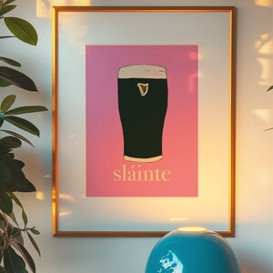 May include: A framed print with a pink background featuring a black and white illustration of a pint of Guinness with the word "sl&aacute;inte" written below it.