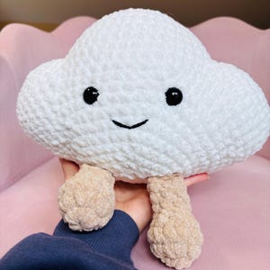 May include: A white plush cloud toy with a soft, textured surface, featuring two black circular eyes and a stitched black smile. The cloud has two dangling, light brown, textured legs attached to the bottom, creating a whimsical character.