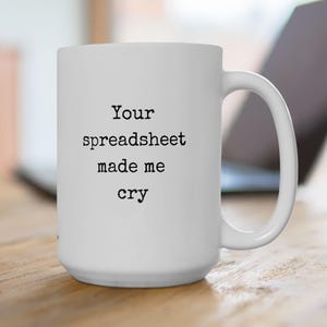 May include: A white ceramic mug with a handle on the right side. The front of the mug features black typewriter-style text that reads: Your spreadsheet made me cry. The mug has a cylindrical shape and a smooth, glossy finish throughout the exterior.