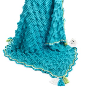 Puede incluir: Una manta de bebé de crochet azul turquesa con un patrón ondulado y ribete verde y blanco. La manta tiene borlas en las esquinas.