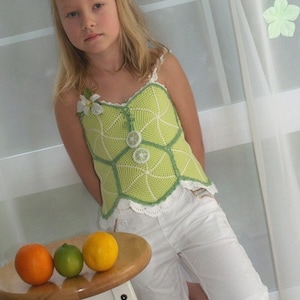 Peut inclure: Une jeune fille portant un haut en crochet vert avec des bordures blanches et un short blanc. Elle se tient à côté d'un tabouret blanc sur lequel se trouvent trois oranges et un citron vert.