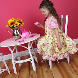 May include: A young girl in a yellow floral dress with a pink crocheted cardigan sits at a white wicker table with a pink teapot and teacups. The table is set on a wooden floor with a pink wall in the background.