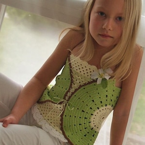 Peut inclure: Une jeune fille portant un haut crocheté vert, marron et blanc avec un accent de fleur blanche. Le haut a un bord festonné et une cravate à la taille. La fille porte un pantalon blanc avec une bordure en dentelle.