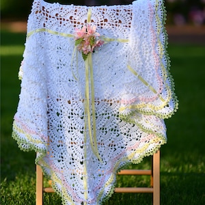 May include: A white crocheted baby blanket with a delicate floral design and a yellow ribbon. The blanket is draped over a wooden chair and has a green, pink, and yellow border.