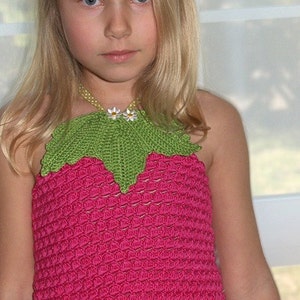 May include: A young girl wearing a pink crocheted top with green leaves and white flowers. The top has a white crocheted trim. The girl has blonde hair and is wearing a white skirt.