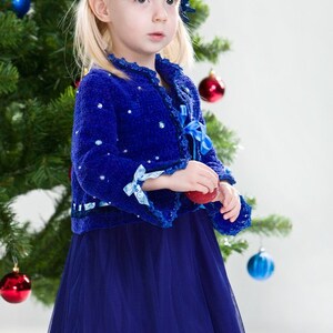 Puede incluir: Una niña pequeña con un cárdigan azul de punto, una falda de tul azul y lazos azules en el pelo. Está sosteniendo un adorno navideño rojo delante de un árbol de Navidad.