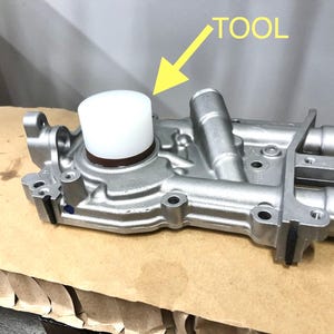 May include: A silver automotive water pump with a white plastic tool on top. The tool is labeled "TOOL" in yellow text.