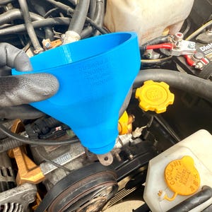 May include: A blue plastic funnel being held over the engine of a car. The funnel has the text "SUBARU Genuine Parts" printed on the side.