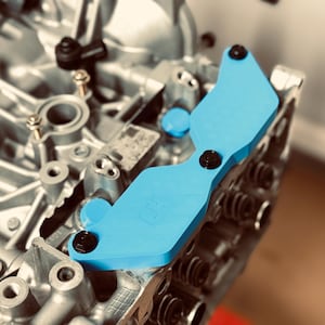 May include: A light blue automotive part with black fasteners is mounted on a silver engine component. The part has a curved shape and the letters "LH" are visible. The background is blurred.