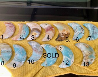 Könnte beinhalten: Eine Sammlung von dreizehn halbmondförmigen Edelstein-Schnitzereien in verschiedenen Farben, darunter Rosa, Blau, Braun und Weiß. Die Schnitzereien sind von 1 bis 13 nummeriert, eine ist mit "SOLD" gekennzeichnet. Die Edelsteine liegen auf einem gelben Tuch.