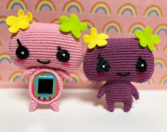 May include: Two crocheted plush toys with smiling faces and large eyes. The pink toy has a yellow flower on its head and a Tamagotchi-like device on its chest. The purple toy has a green flower on its head. Both toys are sitting on a pink and rainbow background.