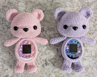 May include: Two crocheted plush teddy bears in pink and purple. Each bear is holding a toy that resembles a Tamagotchi.