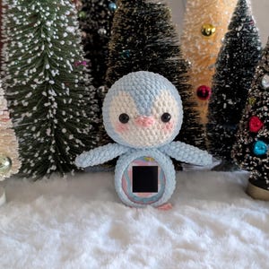 Puede incluir: Un pingüino de ganchillo hecho a mano, azul claro y blanco, con pico y mejillas rosas. El pingüino tiene los brazos extendidos y una pequeña pantalla negra cuadrada en el vientre. Pequeños árboles de Navidad de varios colores y tamaños en el fondo.