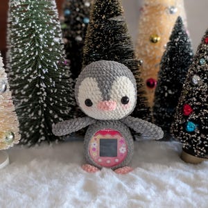 May include: A crocheted penguin toy in gray, white, and pink, with a screen on its belly. The penguin is set against a backdrop of miniature Christmas trees with colorful ornaments, creating a festive holiday scene.