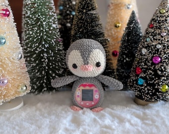 May include: A crocheted penguin toy in grey, white, and pink, with a screen on its belly. The penguin is set against a backdrop of miniature Christmas trees with colourful ornaments, creating a festive holiday scene.