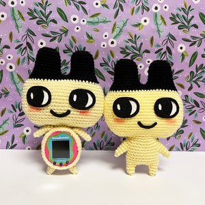 Może przedstawiać: Dwie pluszowe zabawki wykonane na szydełku, przypominające postacie z Tamagotchi. Obie są żółte z czarnymi uszami i oczami. Jedna zabawka trzyma różowe i niebieskie Tamagotchi.