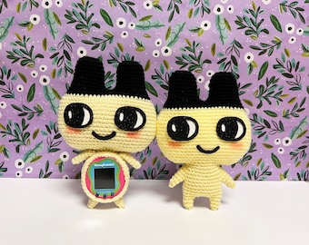 May include: Two crocheted plush toys resembling the Tamagotchi characters. Both are yellow with black ears and eyes. One toy is holding a pink and blue Tamagotchi.