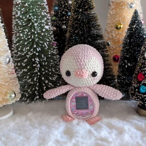 Puede incluir: Un dispositivo con forma de pingüino tejido en rosa y blanco, con una pequeña pantalla, se encuentra frente a un fondo de árboles de Navidad en miniatura. El pingüino tiene ojos negros y un pico y pies rosas. Los árboles están adornados con adornos coloridos.