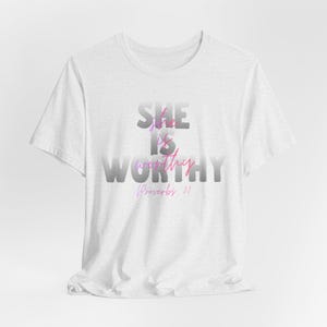 Può includere: T-shirt bianca con la scritta "SHE IS WORTHY" in un gradiente di grigio, rosa e viola. Il testo "Proverbi 31" è stampato sotto. La maglietta è realizzata in un tessuto morbido e di colore chiaro.