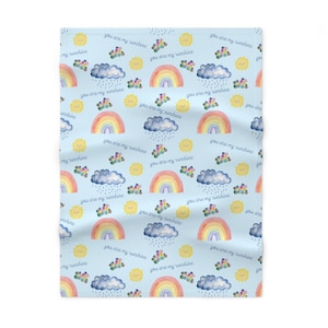 May include: A light blue blanket with a repeating pattern of rainbows, clouds, suns, and floral accents. The words "you are my sunshine" are written across the blanket in a cursive font. The blanket is made of a soft material.