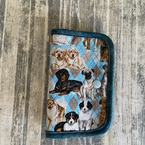 May include: A blue denim fabric wallet with a white button closure. The wallet is decorated with a pattern of various breeds of puppies in a blue and brown plaid design.
