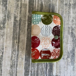 May include: A small, rectangular, green-trimmed fabric pouch with a white background and a colorful pattern of red, orange, and green circles. The circles have different patterns and designs.