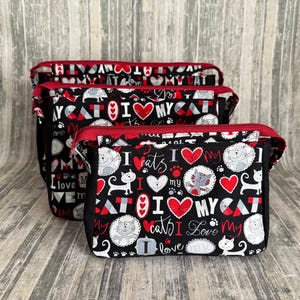 May include: Set of three black zippered pouches with a red trim. The pouches are decorated with a repeating pattern of cats, hearts, and the phrase "I love my cats". The background is a light gray wood.