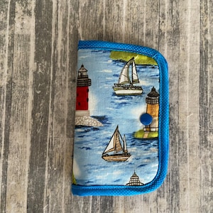 May include: A blue fabric wallet with a nautical theme. The wallet features a lighthouse, sailboats, and a blue button closure.