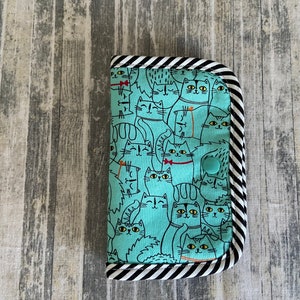 May include: A teal blue fabric wallet with a black and white striped border. The wallet is covered in a pattern of black and white cats with various expressions.