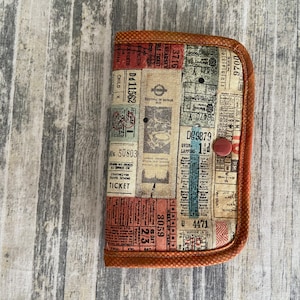 May include: A brown fabric wallet with a vintage design featuring a collage of old train tickets. The wallet has a red button closure and a brown stitched border.