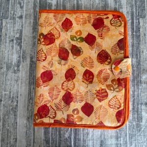 May include: A rectangular fabric folder featuring a repeating pattern of stylized leaves in shades of red, orange, and yellow on a light background. The folder has a solid orange fabric border and a small tab closure secured with an orange snap.