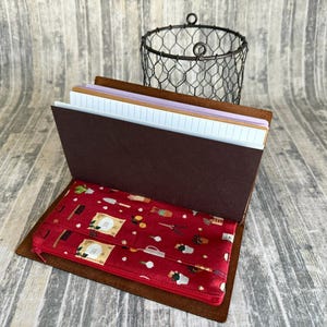 May include: Open brown leather journal with a red patterned interior. The interior features a red zippered pouch with a pattern of kitchen tools and food items. The journal is open to reveal lined and blank pages.