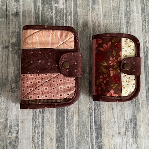 May include: Two quilted fabric zippered pouches, one larger than the other. The larger pouch has a brown, pink, and white patchwork design. The smaller pouch has a brown, white, and pink floral design.