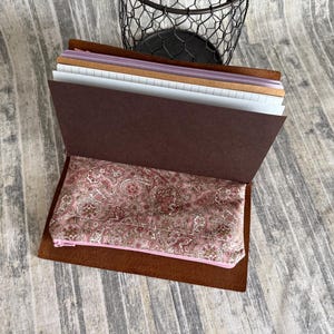 May include: A brown leather journal cover containing multiple paper inserts and a pink fabric zippered pouch with a detailed paisley pattern. The journal is open, revealing white lined pages, a brown cardstock divider, and the patterned pouch.
