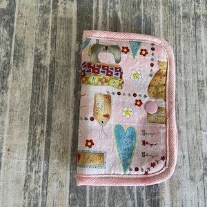 May include: A pink fabric sewing kit with a button closure. The fabric features a sewing themed print with a sewing machine, a cat, and the text "Sewing fills my heart with joy".