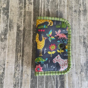 May include: A small grey fabric pouch with a green and yellow trim. The pouch has a colourful pattern of animals and flowers. A black button is visible on the front of the pouch.