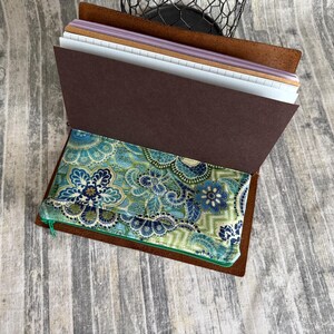 May include: A brown leather journal cover containing multiple paper inserts and a fabric pouch with a blue, green, and white floral paisley pattern. The journal features a dark brown interior flap and a zippered pocket for storage.