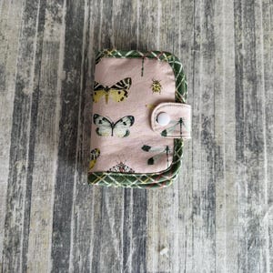 May include: A small, pink fabric wallet with a button closure. The wallet features a pattern of butterflies, dragonflies, and ladybugs. The edges are trimmed with green plaid fabric.