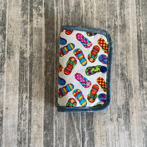 May include: A white fabric wallet with a blue denim trim and a colorful flip-flop pattern. The wallet has a blue button closure.