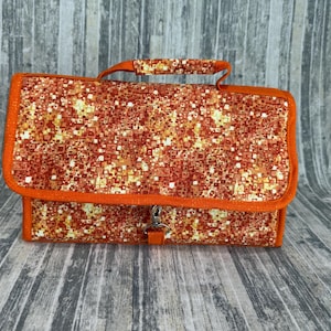 May include: A rectangular fabric organizer bag featuring a vibrant orange, red, and yellow geometric mosaic pattern. The bag includes a top handle, a fold-over flap with orange trim, and a central metal swivel clasp closure on the front.