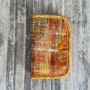May include: A small, rectangular pouch with a zipper closure. The pouch is made of a fabric with a colorful abstract pattern in shades of orange, yellow, and red. The pouch has a brown trim and a small orange button.