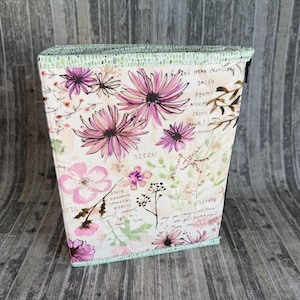 May include: A rectangular fabric container with a floral design. The container features pink and purple flowers, green leaves, and handwritten text on a cream background. The top and bottom edges are trimmed with a light green fabric.