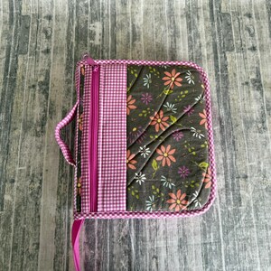 May include: A square, zippered case with a handle and a pink ribbon. The case features a floral pattern on a dark background, a pink and white gingham stripe, and a pink zipper. The edges are trimmed with a pink and white gingham pattern.