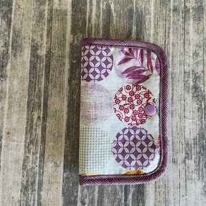 May include: A small purple and white fabric pouch with a button closure. The pouch has a pattern of circles with different designs, including floral and geometric patterns.