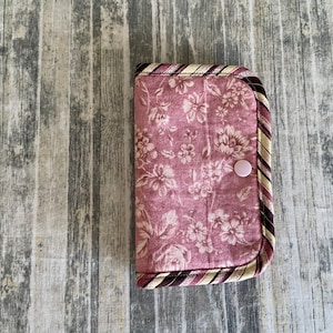 May include: A pink floral fabric wallet with a brown and cream striped trim and a pink button closure.